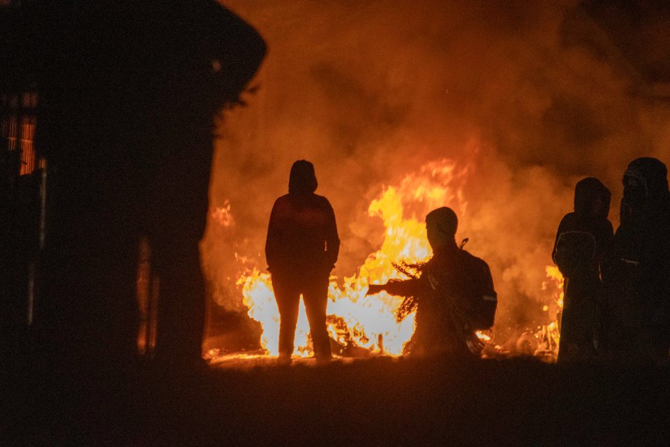 Hooded silhouettes look on as fire engulf the streets during last year riots